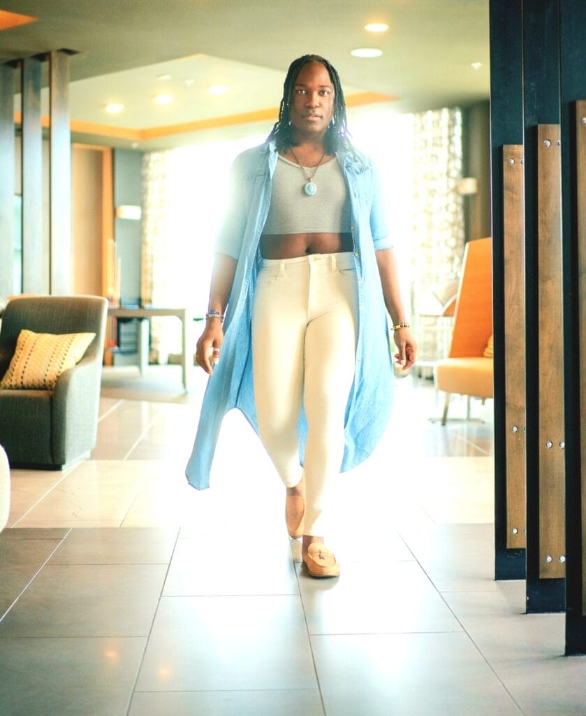 Jourdan Frost walking confidently down a sunlit hallway in stylish attire, featuring a long oversized blue shirt with sleeves rolled up, a grey crop top, and cream bottoms. Modern and serene ambiance.