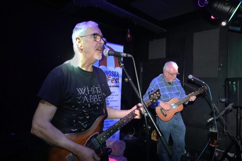 Two musicians, one of which is TG Trouper, performing on a dimly lit stage. One plays guitar and sings into a microphone, wearing a "Whitby Abbey" t-shirt, while the other plays a ukulele. A festival banner is visible in the background, creating a lively and engaging atmosphere.