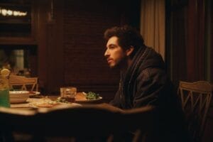Daniel Arison in a dark hoodie sitting alone at a dimly lit dining table, surrounded by plates of food and drinks, conveying a contemplative and pensive mood.