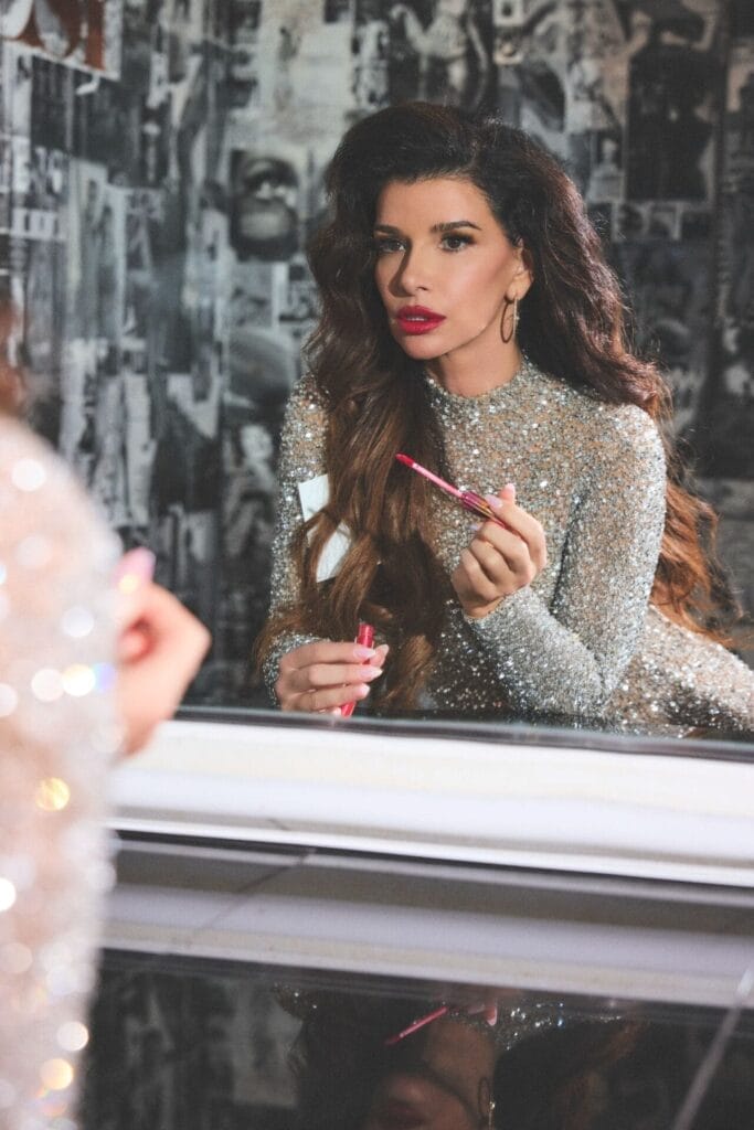 Gina La Piana in a shimmering silver dress applying pink lipstick, gazing into a mirror. Background features black and white collage, conveying glamor.