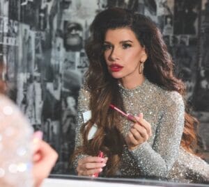 Gina La Piana in a sparkling dress applying lipstick, gazing into a mirror. The setting has a glamorous, vintage feel with a black-and-white photo collage background.