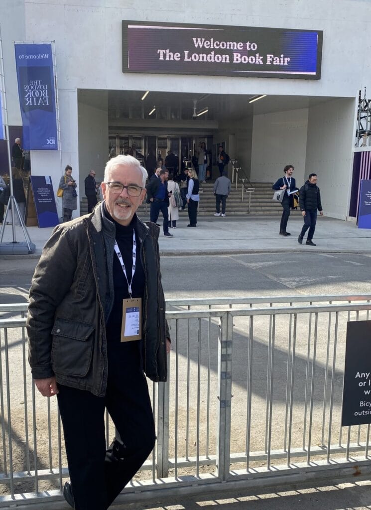TG Trouper standing and smiling in front of the entrance to the London Book Fair. A digital sign welcomes attendees. People walk up steps into the event.