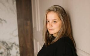 Natalia Kovalchuk with long, light brown hair leaning against a neutral-toned wall, looking calmly at the camera. Marble and wood textures are in the background.