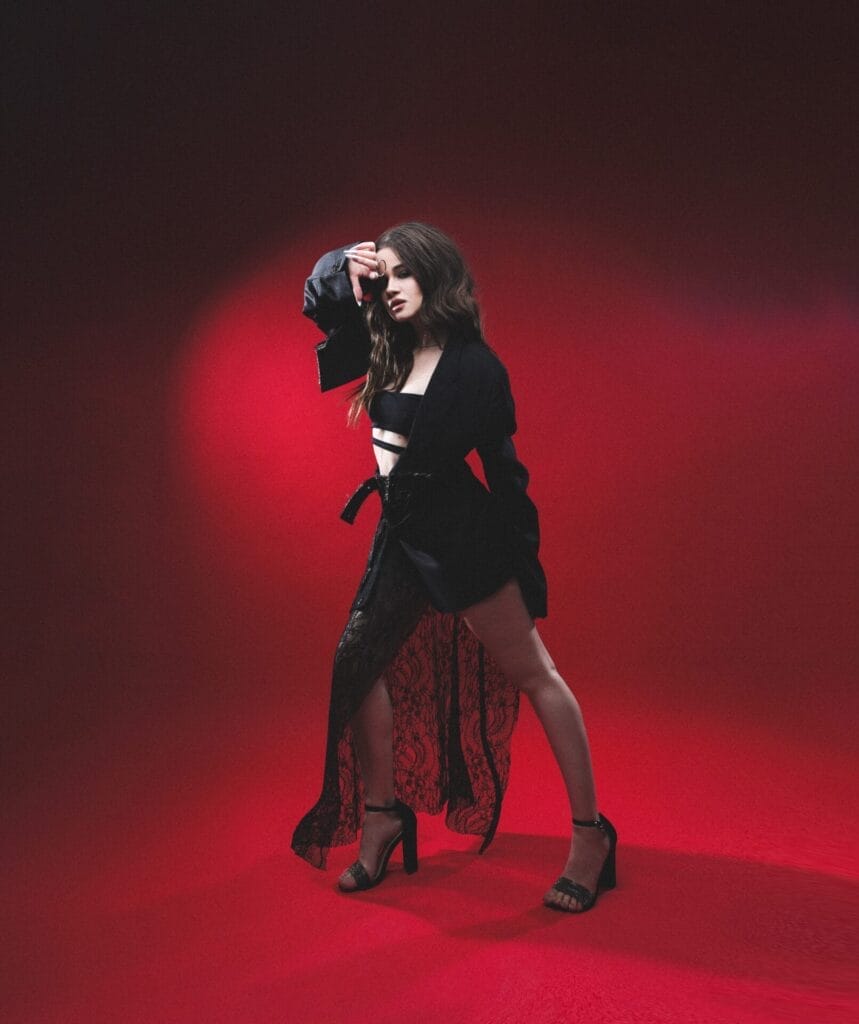 SOWA Opens Up About Embracing Authenticity and Preparing for a New Musical Era 15 SOWA in a stylish black outfit posing confidently against a vibrant red backdrop. She wears a long lace skirt and an open black jacket paired with black high heels, exuding a bold, dramatic tone.