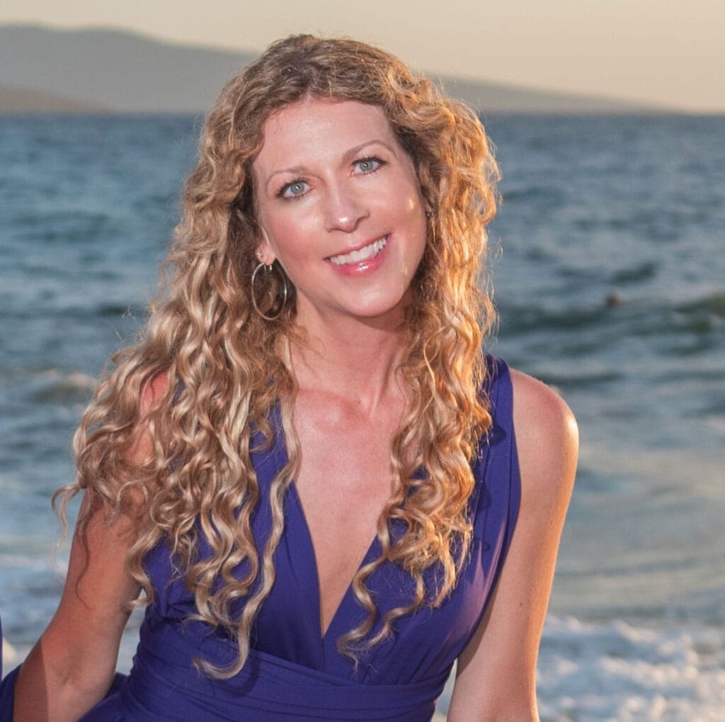Metaphysical Magick: Bestselling Author Megan Mary Crafts Final Autumnal Tale for New Fiction Novel "The Dream Dimensions" 39 Megan Mary with long curly hair smiles warmly at the camera. She's wearing a sleeveless blue dress, standing by the ocean with mountains in the background.