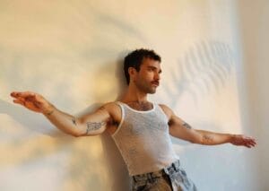 Promotional image for "Off The Ring", which shows Chris Garneau with tattoos wearing a white mesh tank top leaning against a wall with his arms outstretched. Sunlight casts a fern-like shadow, creating a relaxed, artistic vibe.