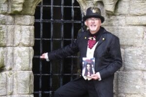 TG Trouper in a black steampunk outfit, with a decorated top hat and glasses, stands in front of a stone wall with iron bar gate at Whitby Abbey, holding his book with a vampire woman's face on the cover, which is titled "Abbey". The atmosphere is mysterious and Gothic.