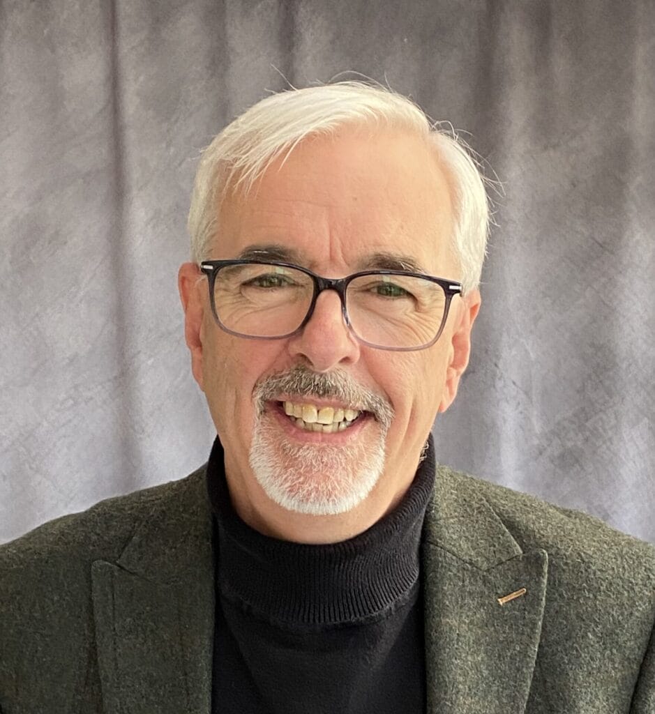 TG Trouper with white hair and glasses smiling warmly at the camera. He wears a dark turtleneck and blazer against a plain grey backdrop.