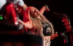 Kelli Baker with long blonde hair standing on stage, eyes closed, arms outstretched, and singing passionately into a microphone. The scene is warmly lit, creating an emotional and vibrant atmosphere.