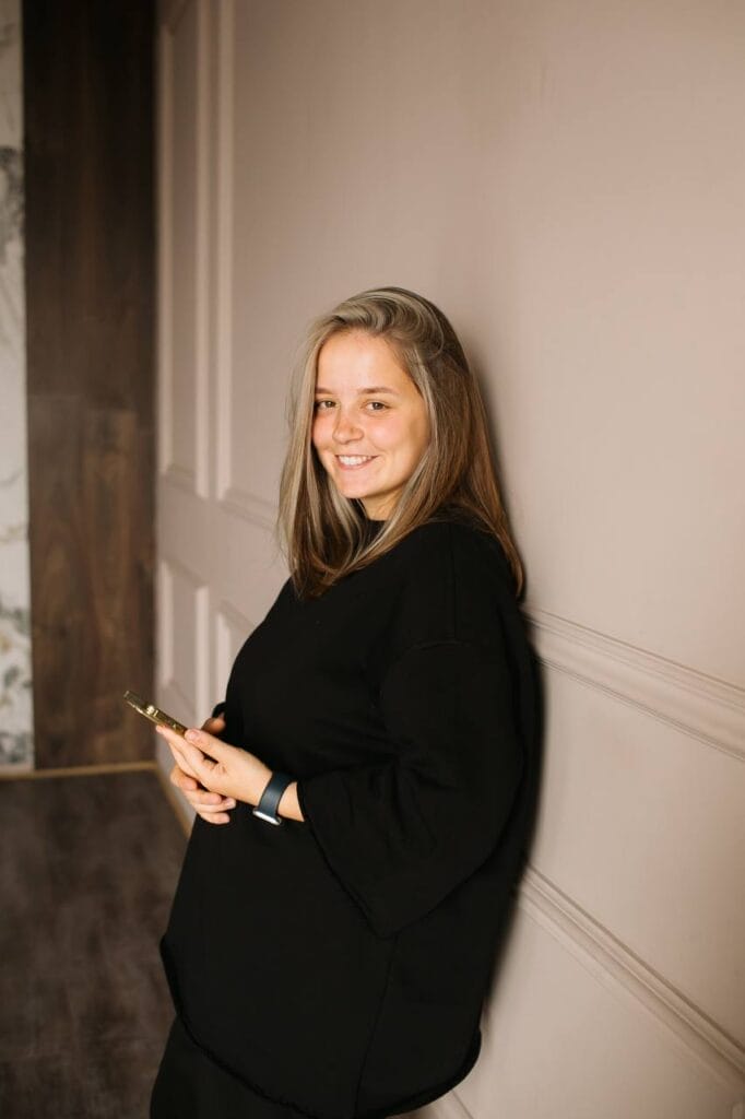 Natalia Kovalchuk Is Breaking the Classroom Walls: How a Ukrainian Educator Is Making Learning More Inclusive Through Creativity and Social Media 14 Natalia Kovalchuk with long hair, wearing a black outfit and holding a phone, leaning against a beige wall, smiling warmly. The setting is softly lit and elegant.