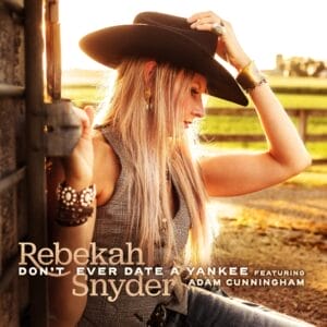 Official single cover artwork for "Don't Ever Date a Yankee" which shows Rebekah Snyder in a cowboy hat leaning against a wooden fence, bathed in warm golden light. Her expression is thoughtful. Text overlay at the bottom reads: "Rebekah Snyder - Don't Ever Date a Yankee featuring Adam Cunningham."