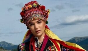 Galina Denisiuk wearing a vibrant red traditional Ukrainian headdress adorned with colourful beads and pom-poms, set against a mountainous backdrop under a cloudy sky.