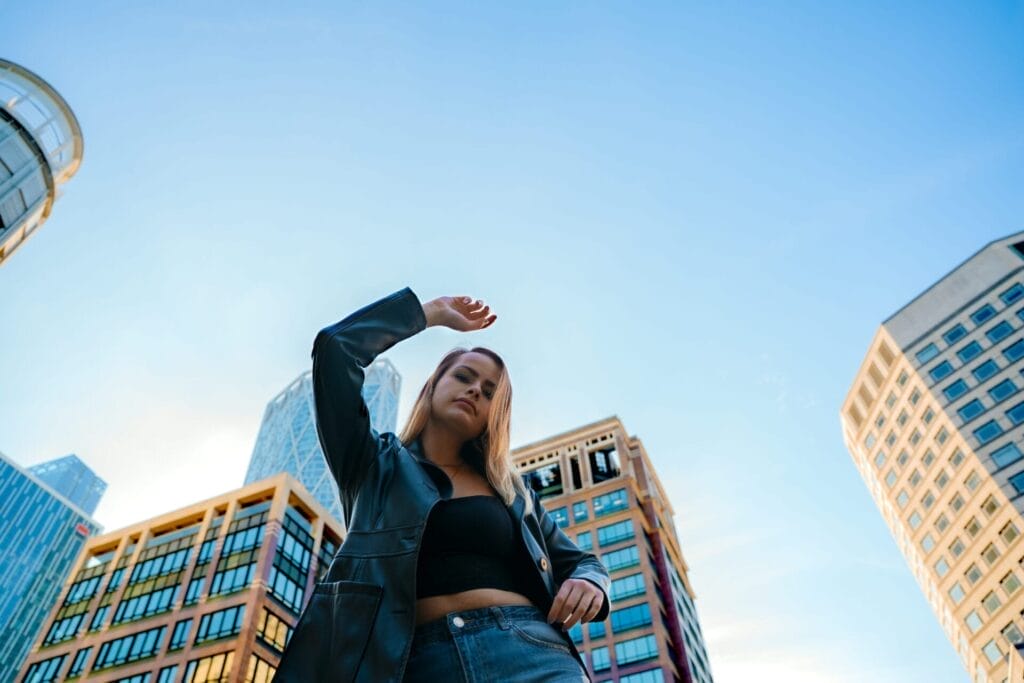 ELUNIA Pushes the Boundaries of Immersive Pop with Emotional New Track “Strobe Lights” 14 ELUNIA looking down at the camera, with an arm raised above her head and the other on her hip. She's wearing a leather jacket over a black vest top, and she's surrounded by high-rise buildings and the sky acts as the background.
