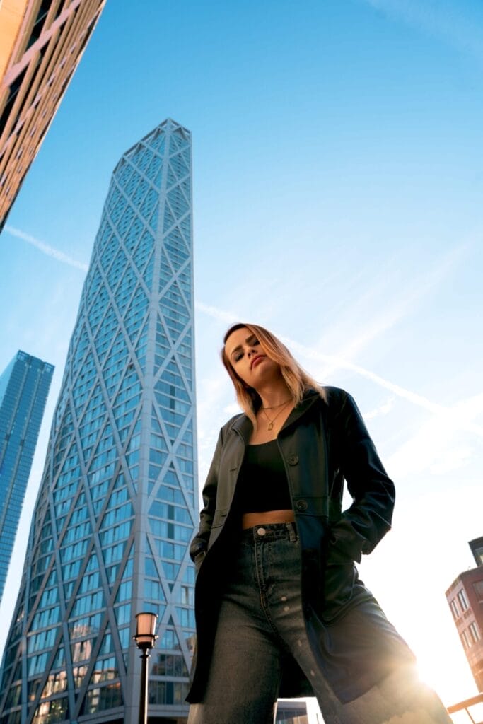 ELUNIA Pushes the Boundaries of Immersive Pop with Emotional New Track “Strobe Lights” 15 ELUNIA posing in front of a high-rise building with her hands in the pockets of her black leather jacket which she's paired with a black vest top and a pair of jeans.
