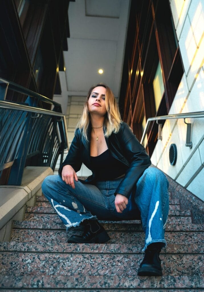 ELUNIA Pushes the Boundaries of Immersive Pop with Emotional New Track “Strobe Lights” 13 ELUNIA sitting on a set of stone steps, wearing a black leather jacket over a black vest top, a pair of jeans with white paint graphic detail on the ends, and a pair of black Chelsea boots.