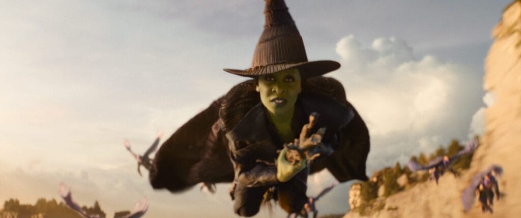 Cynthia Erivo as Elphaba, a green-skinned witch wearing a pointed hat and a dark cloak, flying on a broomstick against a cloudy sky, with flying monkeys in the background.