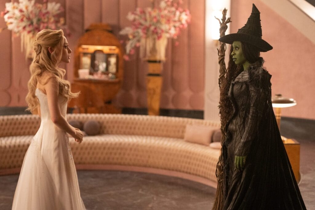 Ariana Grande as Glinda and Cynthia Erivo as Elphaba face each other in an elegant, pink-hued room. The former is in a pale pink gown with long blonde hair, and the latter is a green-skinned witch in dark attire.