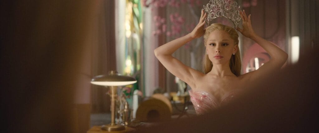 Ariana Grande in a pink dress standing before a mirror in an elegant room, placing a large silver crown on her head. The atmosphere is regal and reflective.