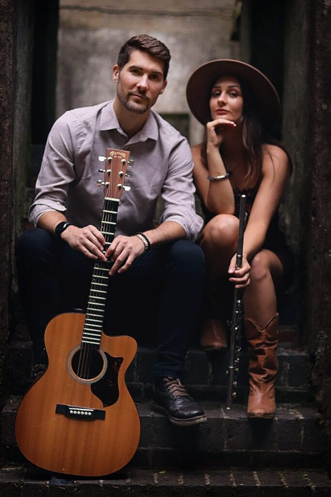 Briggs & Stark posing together with Briggs holding a guitar and Stark holding a flute while sitting on some stone steps. Briggs wears a light shirt, and Stark is wearing a hat and boots and gazing thoughtfully.