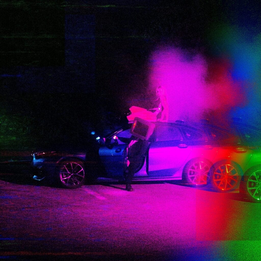 Official single cover artwork for "When I Think About Love" by JVK which shows a surreal scene with a person wearing a TV on their head sitting a car with the door open, while Jo Krieger sits on the roof, under colourful, smoky neon lights. The atmosphere is mysterious and vibrant.
