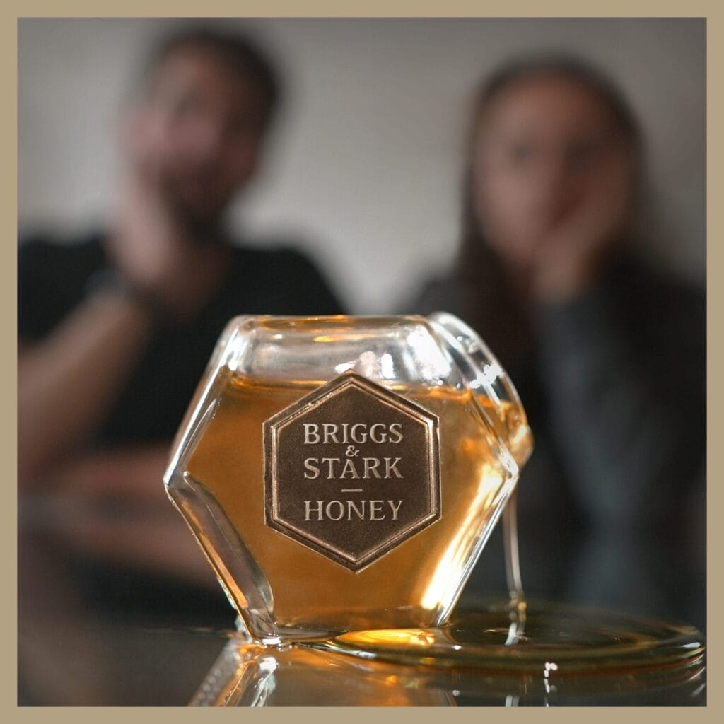 Official single cover artwork for "Honey" which sees a hexagonal glass jar of "Briggs & Stark Honey" spilling on a reflective surface in the foreground while Briggs & Stark can be seen in the blurred background, creating a warm, contemplative scene.