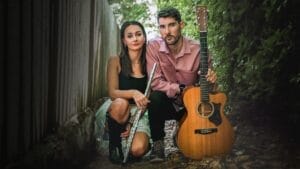 Briggs & Stark posing together with Stark holding a flute and Briggs holding a guitar. They're crouching together on a leafy path, framed by a wooden fence and greenery, conveying a calm, artistic vibe.