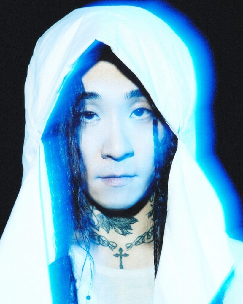 Bryan Chase with long hair and tattoos, illuminated by a blue glow, wearing a white hood. The expression is calm, and the tone is ethereal and mysterious.