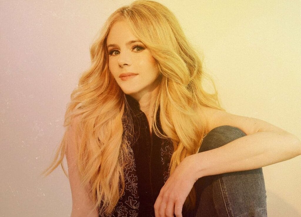 Country-Pop Star Brooke Moriber Shares Bold New Single “Your Jesus” Highlighting Personal Redemption 27 Brooke Moriber with long, wavy, blonde hair sits against a pale background, wearing a black top and jeans. She gazes softly into the camera, conveying a calm mood.
