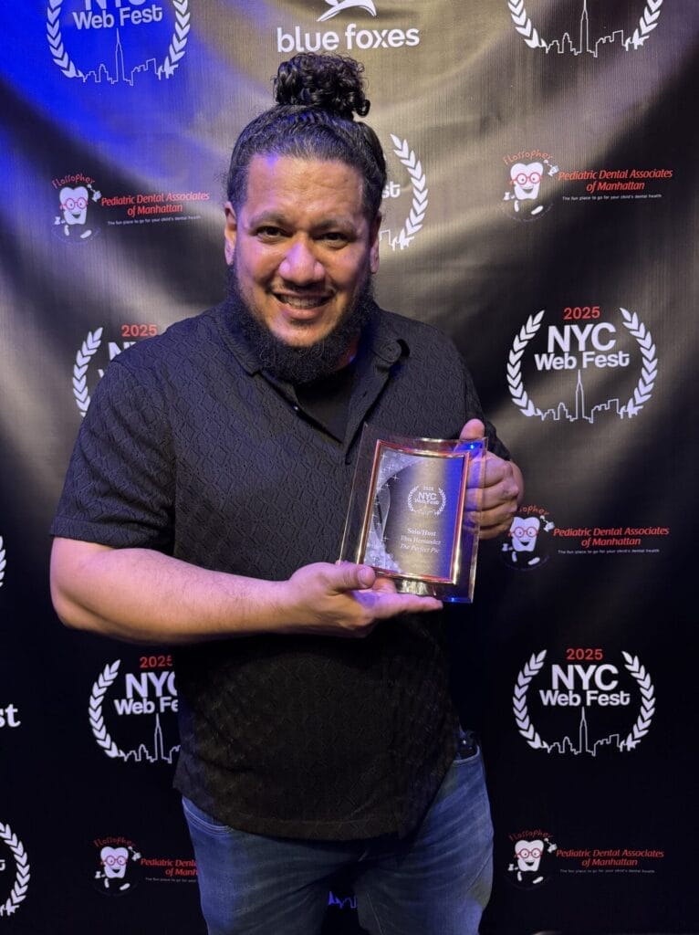 Ebis Hernandez smiling and holding an award at the 2025 NYC Web Fest. The backdrop features sponsor logos and festival branding, creating a celebratory atmosphere.
