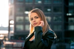 ELUNIA looking side-on to the camera with her blonde hair flowing past her shoulders as she raises a hand to her cheek. She's wearing a leather jacket over a black vest top and there's a building behind her that's blurred.