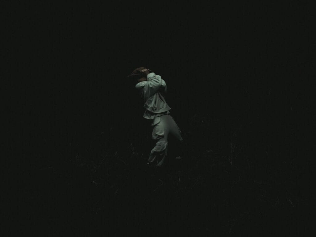 NF in loose, flowing white clothes with a burlap sack over his head standing in a dark, shadowy environment with arms raised to their head, conveying a sense of solitude and mystery.