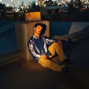 Chris Garneau in a shiny blue shirt and yellow bottoms sits against a rooftop structure at night. City lights twinkle in the blurred background, creating a contemplative mood.