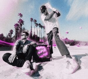 MOVIESTARZ in a vibrant beach scene with palm trees. One member of the duo sits beside a glowing TV, while the other swings a baseball bat. Energetic and playful vibe.