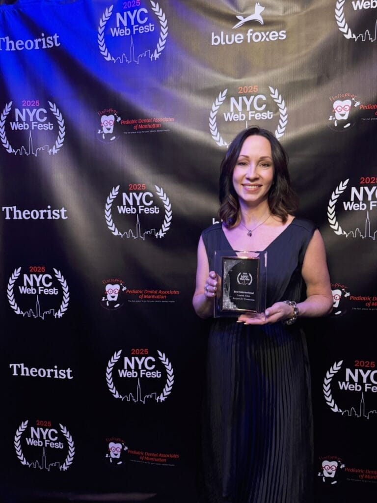 Leanne Allen in a black dress smiles while holding an award in front of a NYC Web Fest backdrop with sponsor logos. The atmosphere is celebratory.