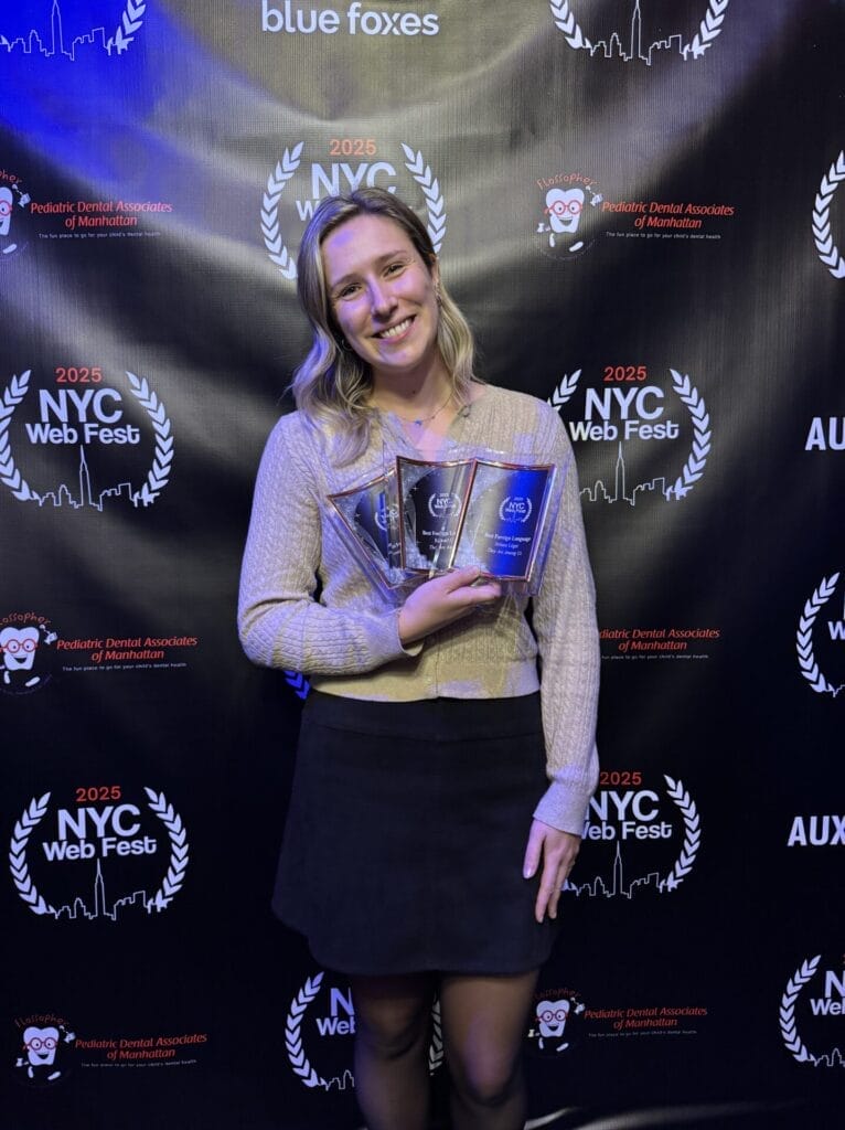 Véronique Paquette-Corriveau smiling and holding three awards at the 2025 NYC Web Fest. She stands against a branded backdrop, expressing pride and achievement.