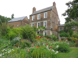 Crook Hall Gardens where a charming three-story stone house with white framed windows can be seen surrounded by a lush, vibrant garden blooming with colorful flowers and greenery.