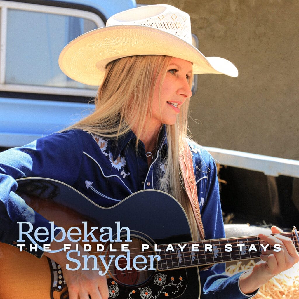 Official single cover artwork for "The Fiddle Player Stays" which shows Rebekah Snyder in a cowboy hat playing a guitar outdoors, wearing a blue embroidered shirt. Text of artist name and song title overlays the image. Energetic mood.