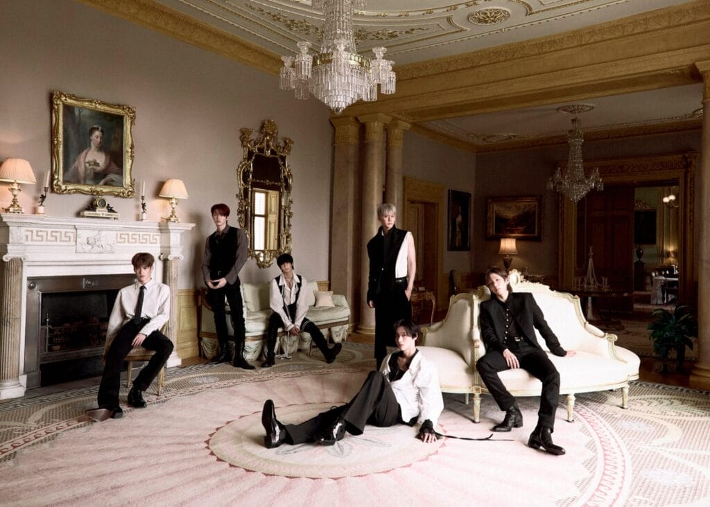 The six band members of RIIZE in elegant black and white outfits posing in a luxurious room with ornate columns, chandeliers, and vintage décor, conveying a refined, classic atmosphere.