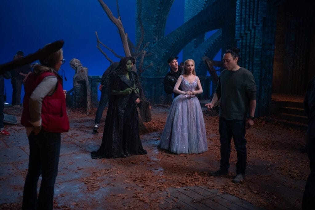 Behind-the-scenes image on the Wicked: For Good set with a fantasy theme. Ariana Grande and Cynthia Erivo are in detailed costumes, including a Erivo in black and a Grande in a sparkling gown, conversing with crew members in front of blue screens and faux forest backdrop. Atmosphere is creative and collaborative.
