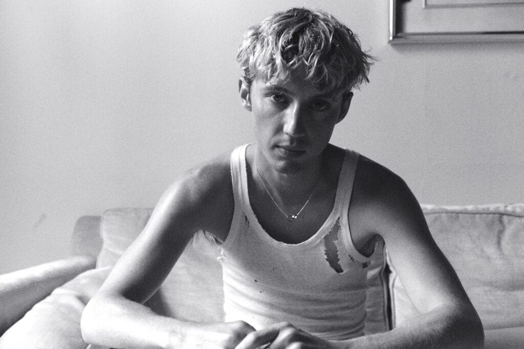 Grayscale image of Troye Sivan in a torn tank top sitting on a couch, with a thoughtful expression.