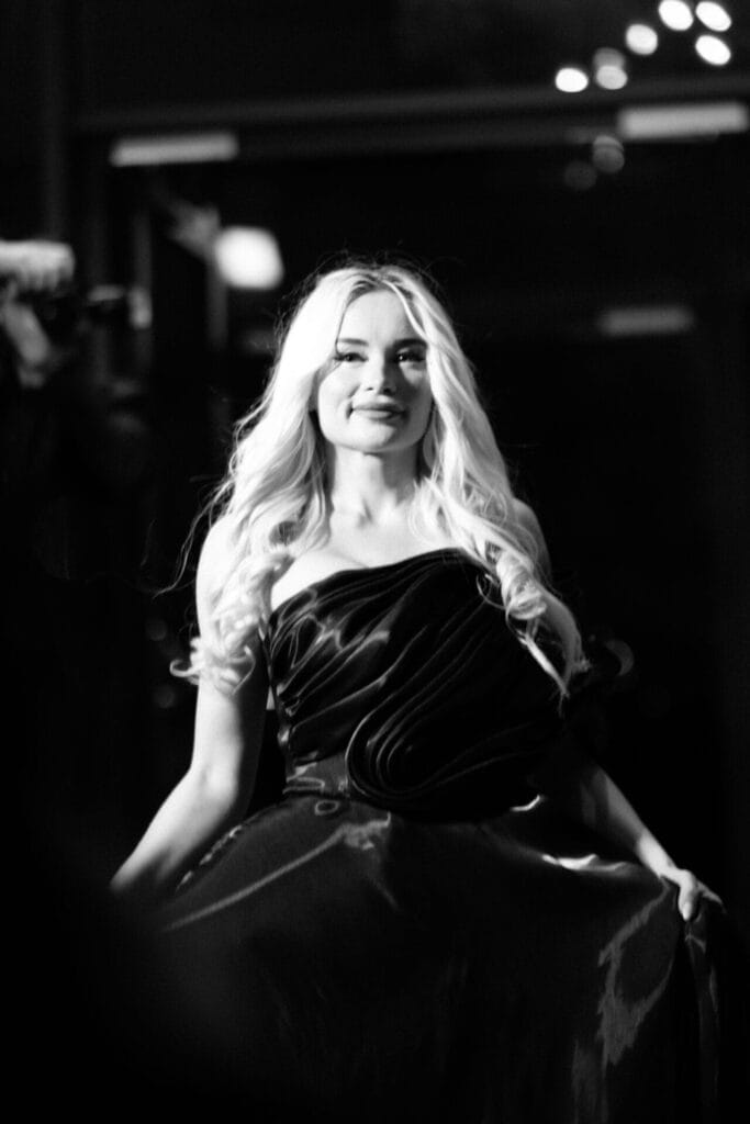 Black and white photo which shows ANGELA with long blonde hair smiling confidently, wearing a flowing, strapless black gown. The black and white setting adds a classic, elegant tone.
