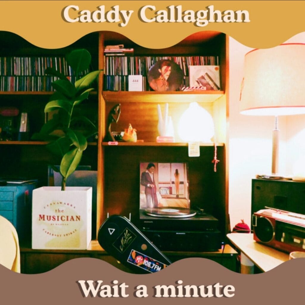 Official single cover artwork for "Wait A Minute" by Caddy Callaghan which shows a cosy room with a wooden shelf, vinyl records, and a lit lamp. Plant and decorative items add warmth. The artist name is at the top with the song's title at the bottom, both in white text.
