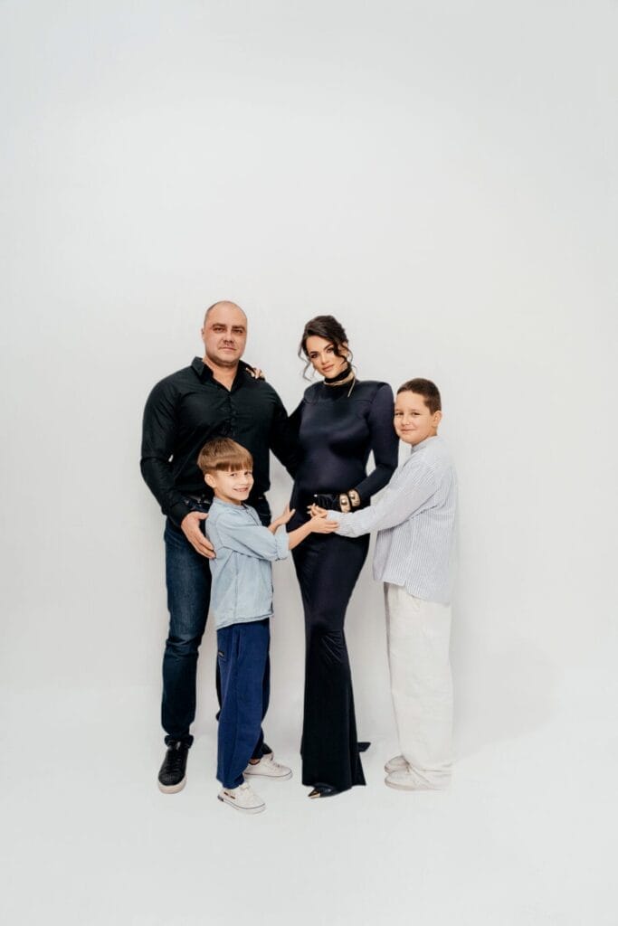 Galina Denisiuk and her family posing for a family portrait showing a smiling mother and father with two young boys, one wearing a blue shirt and the other in white. They stand closely together, touching the mother's baby bump, with a white background, conveying warmth and togetherness.