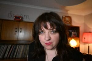 Caddy Callaghan with long dark hair and a nose piercing in a cosy room with vinyl records, warm lighting, and the word "dream" on the wall; calm atmosphere.