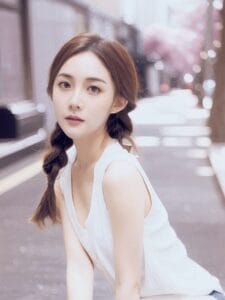 Liz Luceris with braided hair and a white sleeveless top sits on an urban street. Soft lighting and blurred pink trees create a serene, dreamy atmosphere.