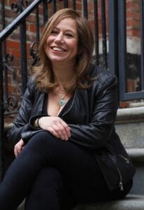 Alyson Faith in a leather jacket sitting on stone steps, smiling warmly. Brick wall and metal railing in the background. Casual and cheerful atmosphere.