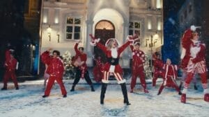 Still from Kylie Minogue's "XMAS" music video which shows the Australian superstar performing with her dance group in a snowy, lit-up town square, wearing red and white Christmas costumes, exuding joy and holiday spirit.