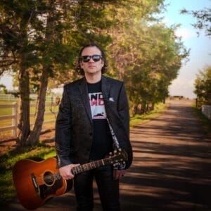 Jeremy Climer (Mountain Climer) in sunglasses and a patterned jacket is holding a guitar on a sunlit tree-lined path. The scene conveys a relaxed and cool vibe.