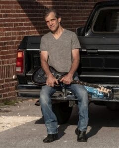 Jordan Seven in a grey t-shirt and jeans sitting on a truck's tailgate holding a guitar. He's smiling slightly, with a relaxed ambiance against a brick wall.