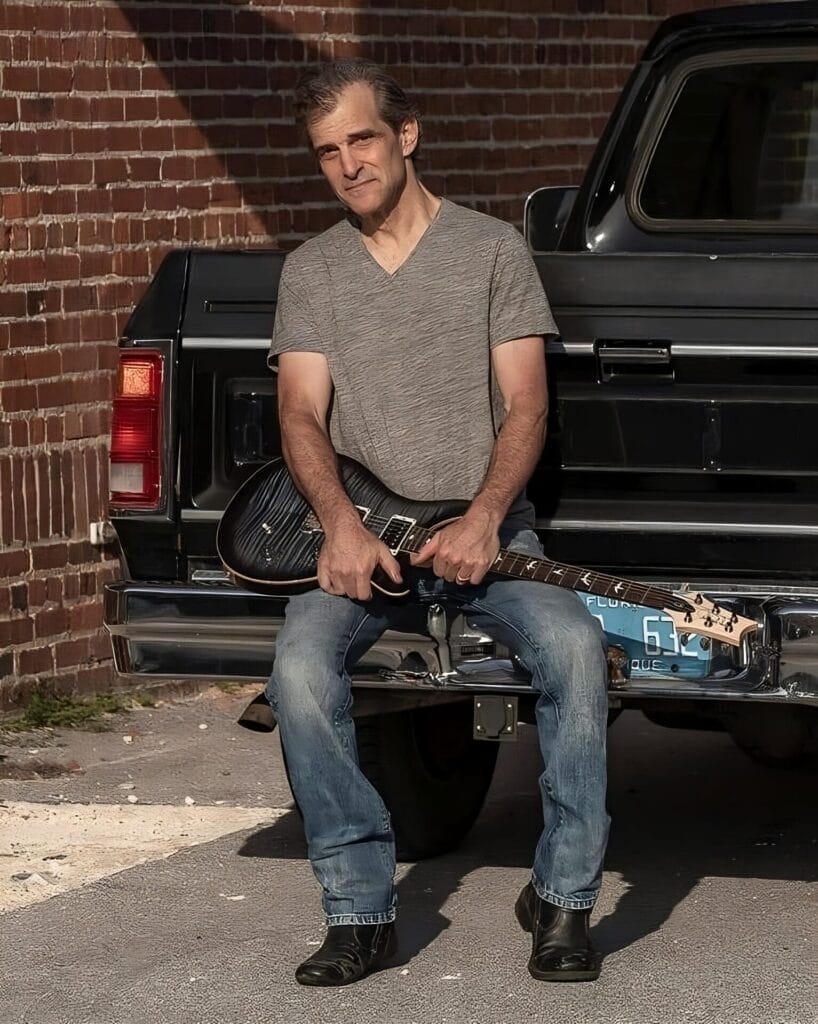 Jordan Seven in a grey t-shirt and jeans sitting on a truck's tailgate holding a guitar. He's smiling slightly, with a relaxed ambiance against a brick wall.